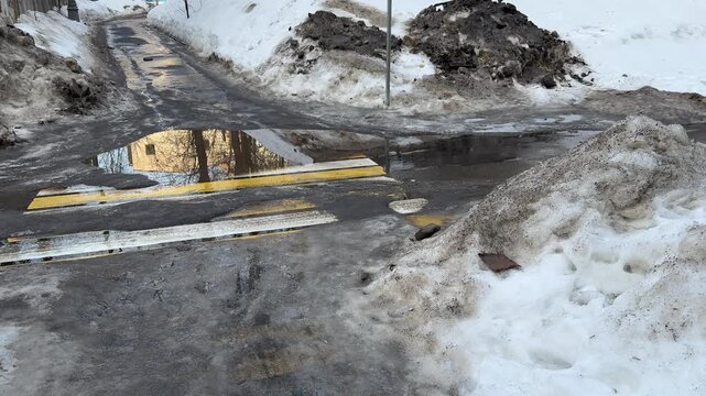 Spring urban scene with melting snow, muddy road, puddles, slushy icy snow, seasonal transition, wet and dirty surface, urban outdoor environment, reflecting sky and surroundings