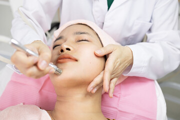 Naklejka premium Young woman relaxes during facial treatment. Professional hands use gentle device on her forehead. Her eyes are closed, showing calm and trust. Soft pink towel wraps her head for comfort 