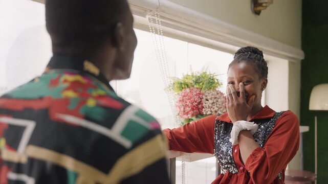 Black woman smiling by window, playful tone while leaning on frame and chatting with man in patterned shirt, bubbly laugh, hand covering mouth at compliment, warm sunlight, affectionate date energy,