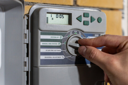 Hand turning selector dial on irrigation controller panel, adjusting automatic watering system settings on a brick wall.