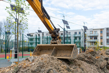Close view of excavator arm and metal bucket resting over excavated soil, with residential buildings, solar lamps and fenced sports area in background, showing urban construction and groundworks. © Volha