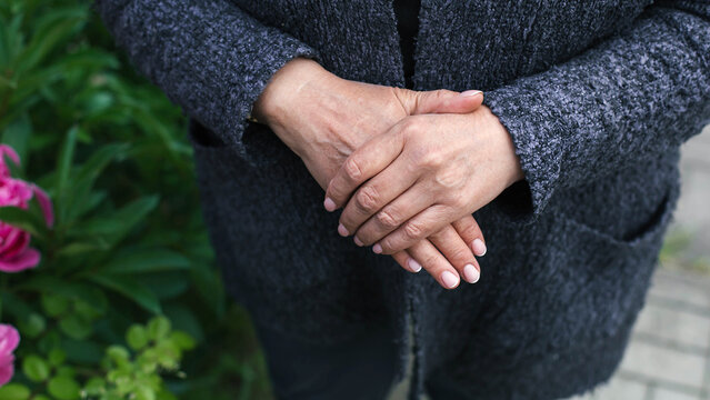 Close up elderly woman hands gently clasped over textured gray coat outdoors, wrinkled skin manicured nails visible, conveying calmness care aging dignity retirement 