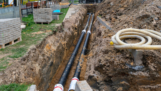 Open construction trench with black utility pipes, coiled protective conduit, paving blocks and excavated soil, showing underground infrastructure installation and urban road repair worksite.
