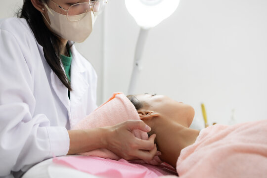 Young woman relaxes during facial treatment. Professional hands gently cleanse her eyes. Soft pink towels frame her serene expression. Relaxed posture suggests comfort and care
