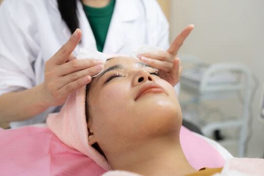 Young woman relaxes during facial treatment. Professional hands gently cleanse her eyes. Soft pink towels frame her serene expression. Relaxed posture suggests comfort and care
