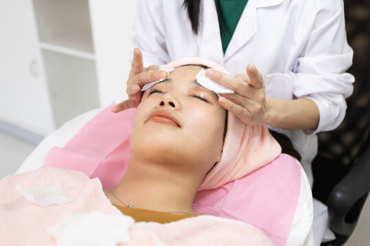 Young woman relaxes during facial treatment. Professional hands gently cleanse her eyes. Soft pink towels frame her serene expression. Relaxed posture suggests comfort and care
