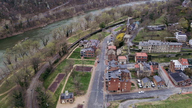Harpers Ferry river curve frames church ridge, clustered brick buildings, rail alignment along Potomac corridor. Mixed roof colors, early leaf-out, hillside shadows create soft spring texture, WV