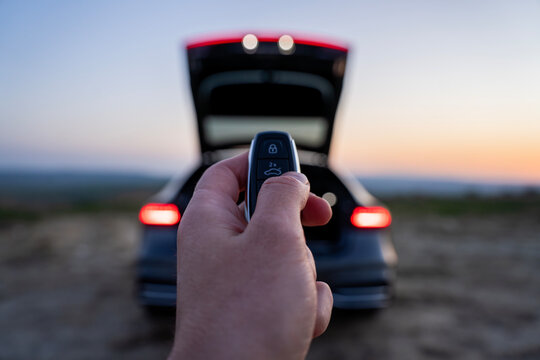 Hand pressing trunk button on car key fob in front of vehicle with open trunk and illuminated rear lights at dusk.