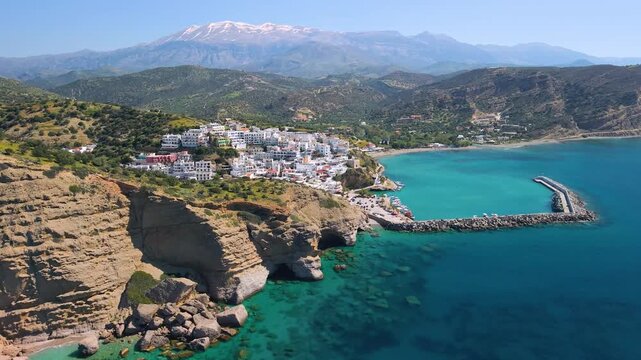 Agia Galini Crete Greece aerial panorama of white village and orange sandstone cliffs, turquoise bay with sea caves, snow capped mountains background on Greek island, spring travel destination