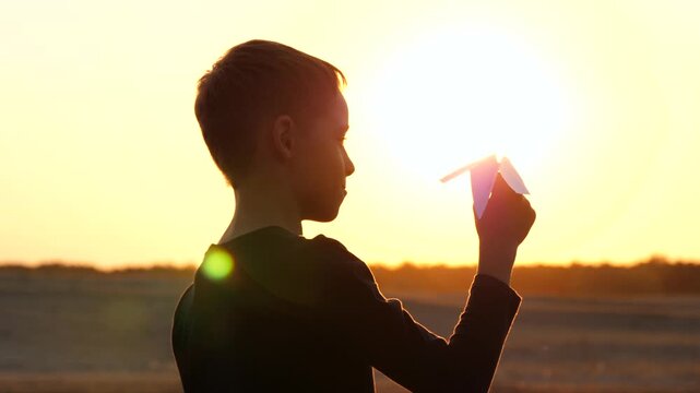 Child playing with paper airplane at sunset, holding it towards sky in warm golden light. Dreams, imagination and future aspirations, creativity and inspiration in happy childhood, outdoor lifestyle.