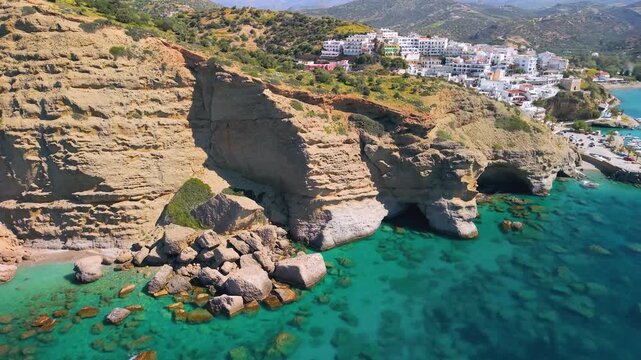 Aerial view of Agia Galini, South Crete. 4-layer panorama: turquoise sea with caves, yellow sandstone cliffs, white village on hills and snowy mountains on background. Scenic Greek landscape, 4k.