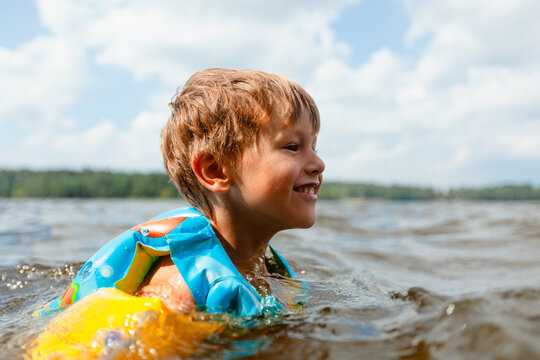 Boy swims in a lake wearing life jacket with arms outstretched on a sunny summer day. Child relaxing on the water surface in a peaceful pose. Kid enjoys safe outdoor recreation