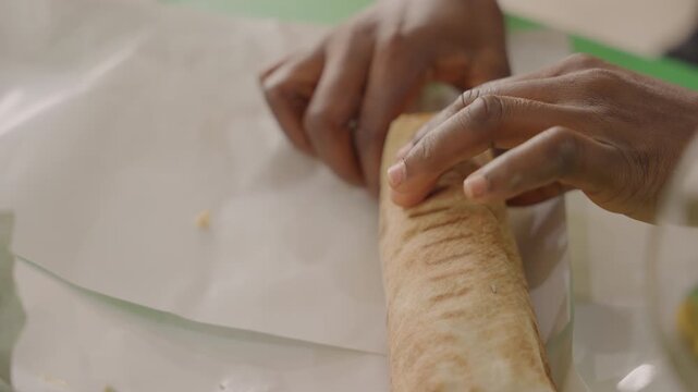Black hands rolling tortilla wrap during outdoor picnic date, closeup of partner assembling warm shawarma wrap on paper with green cutting board, savory chicken and fresh lettuce falling, intimate