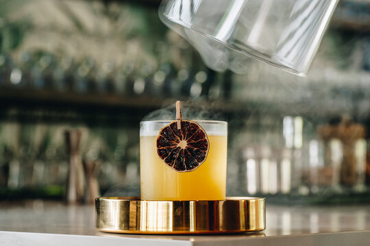 Smoky, flavorful cocktail recipe. A bartender pours smoke over a cocktail with a dried fruit garnish in a bar at night. The setup shows skill and care.