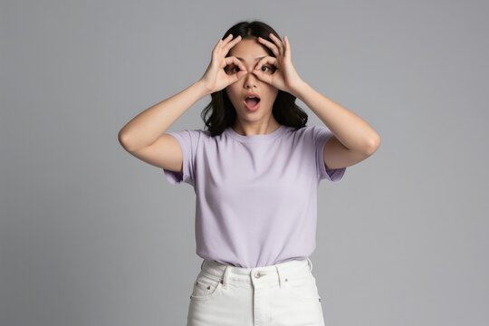 Young Asian woman makes binoculars gesture over eyes. Girl expresses surprise and curiosity with open mouth, looking forward. She wears purple t-shirt and white pants against gray backdrop.