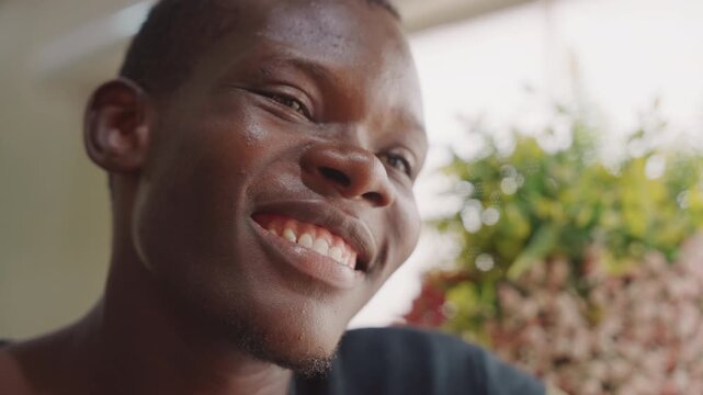 Black man smiling by sunlit window, candid closeup showing joyful expression, subtle laughter, indoor plants and warm ambient light creating cozy romantic atmosphere during intimate conversation