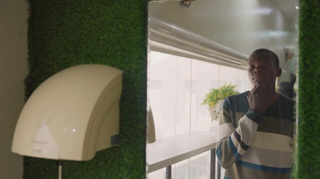 Man by window preparing for date, striped sweater, hand to face, soft sunlight through blinds, greenery wall, warm indoor tones, intimate nervous anticipation before romantic meeting