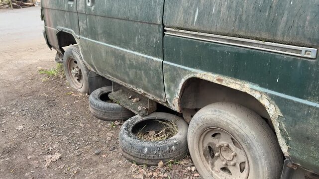 Abandoned green minibus with flat tire, rusted panels and moss-covered windows punctured spare wheels strewn on gravel parking