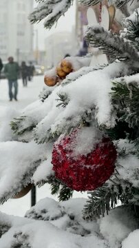 Close up of a red sparkly Christmas bauble on a pine branch covered with fresh snow. People walking on a city street in the background