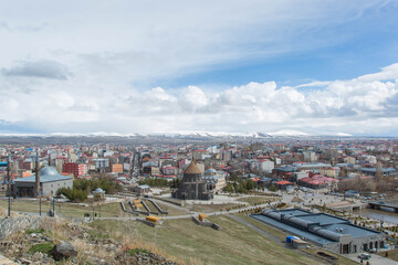 Fototapeta premium Panoramic view of Kars city with historic stone mosque and cathedral buildings, urban landscape with colorful houses and distant snow covered mountains under dramatic cloudy sky, travel 
