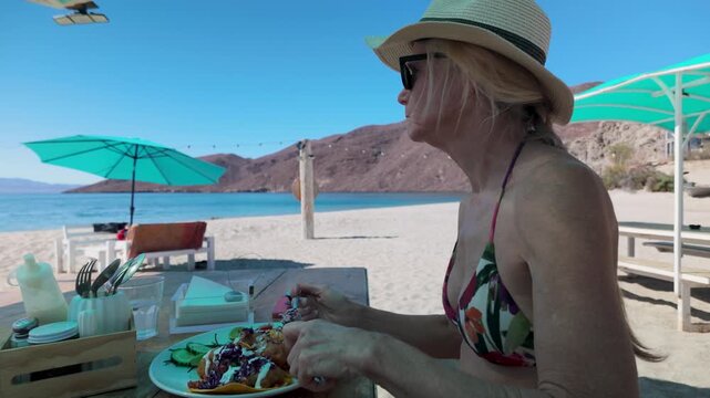 Mature woman eats shrimp tacos at a taco stand on Playa Blanca beach in Baja California on a sunny day.