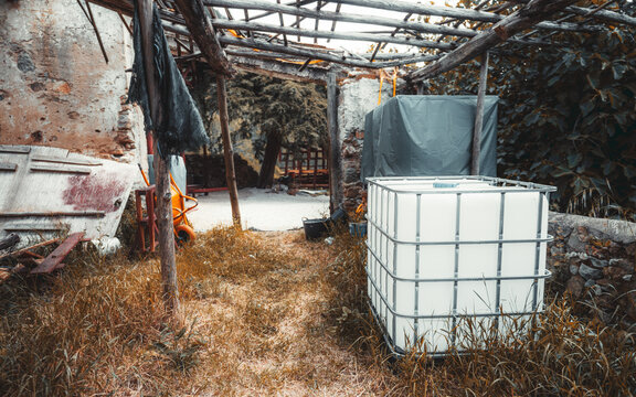 Abandoned rustic courtyard with overgrown grass, wooden roof beams, old stone walls and a white industrial water tank in rural decay setting