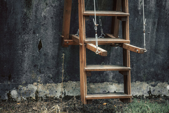Hanging wooden ladder or suspended rustic swing structure with metal cables against dark weathered concrete wall in abandoned outdoor yard