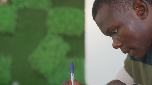 Black man focused on writing closeup profile against moss wall, pen poised over notebook, serious expression preparing vow draft or promise note, quiet cafe interior, concentrated mood with deliberate