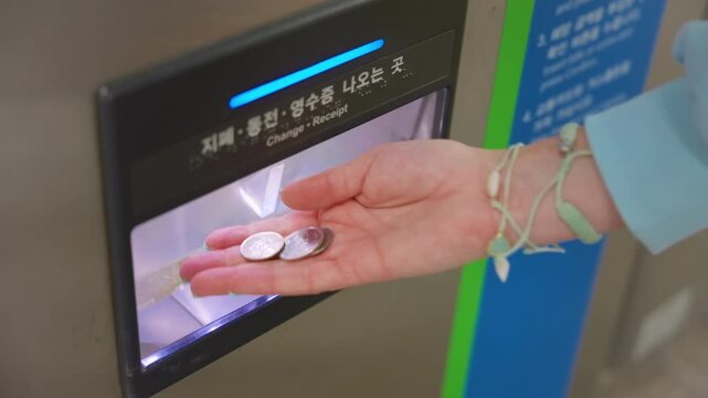 Close-up footage of a woman with mint green manicured nails collecting coins from a Korean subway ticket machine change dispenser, wearing light blue sweater and delicate wrist bracelets.