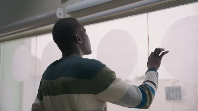Man overlooking street through patterned blind. Back view in striped sweater, soft daylight and muted city skyline beyond glass, hand raised to veil, distant gaze suggesting waiting or missed date,