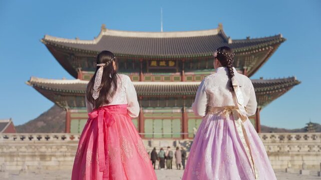 Two women in elegant traditional Korean hanbok dresses with pink and lavender skirts admire the majestic Gyeongbokgung Palace under a clear blue sky in Seoul, tourists visible in the distance.