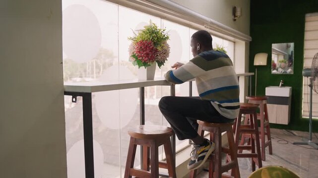 Man checking phone at window counter. Seated man in striped sweater taps phone, bouquet nearby, soft patterned curtain backlight, subtle smile after incoming message, modern green wall interior, warm