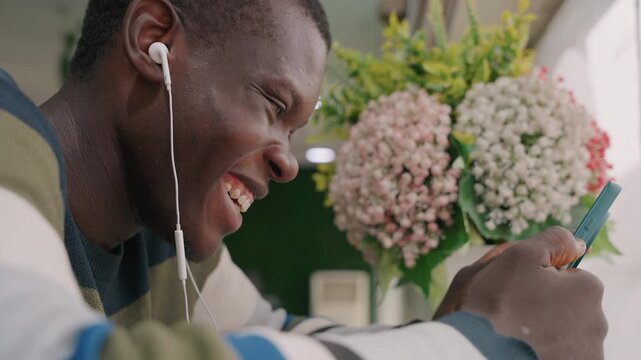 Black man smiling at phone during cozy cafe date with earphones, hydrangea bouquet blurred behind, warm sunlight, affectionate texts and genuine laughter creating intimate modern connection