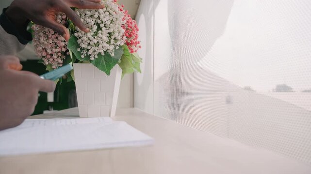 Black man florist checking note by window, arranging hydrangea bouquet with careful hands, pen and paper on desk, phone message open, soft daylight filtering through curtain, focused calm
