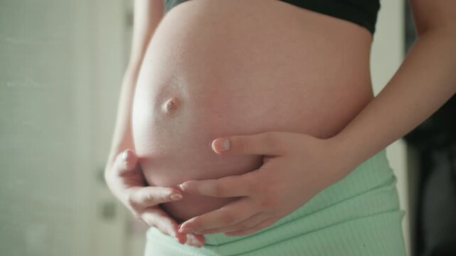 Pregnant caucasian belly with hands gently cradling skin, closeup on navel and subtle texture, mint shorts waistband visible, soft natural light highlighting softness and warmth, nurturing touch