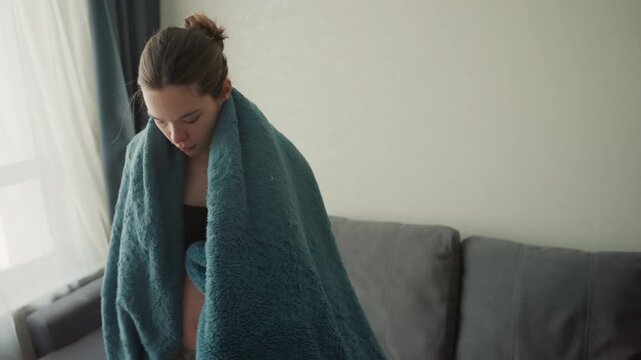 Pregnant caucasian woman wrapping blue blanket over bare belly by window adjusting bra and loungewear settling into sofa warm soft throw reflective tender mood slow breathing quiet evening routine