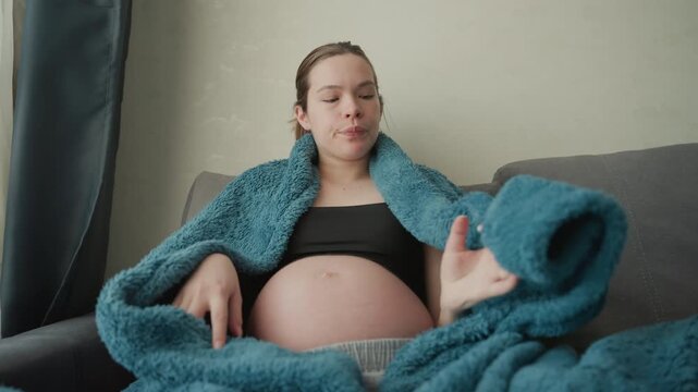 Pregnant white woman resting on sofa wrapped in teal blanket, gentle hand on exposed belly, soft natural light through window, quiet maternity moment, contemplative mood, third trimester rest
