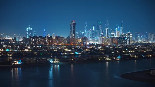 Dubai cityscape glows at night with illuminated skyscrapers of Marina district rising behind waterfront luxury villas. City lights reflect on calm water creating stunning urban panorama.
