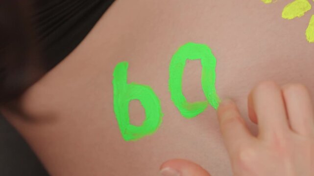 Neon paint on pregnant belly with hand drawing circular figures and sun motif near bra edge, bright green pigment on warm skin, closeup announcement vibe with tactile marking, joyful expectant mood