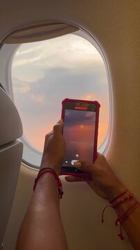 Woman filming sunset through airplane window with smartphone at golden hour