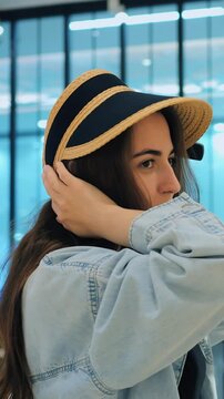 Woman adjusts straw visor reflection. Woman perfects straw visor fit with store display lighting. Fashion enthusiast finetunes straw hat fit while observing herself in store window mirror