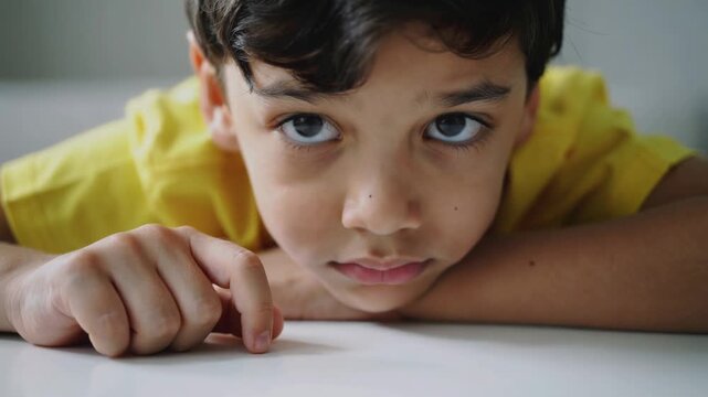 Young boy in yellow t-shirt resting chin on hands looking at camera in close up shot