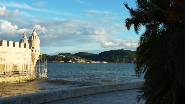 A historic tower stands beside a body of blue water with hills in the background under a cloudy sky. A palm tree provides a foreground element. Belem, Lisbon, Portugal