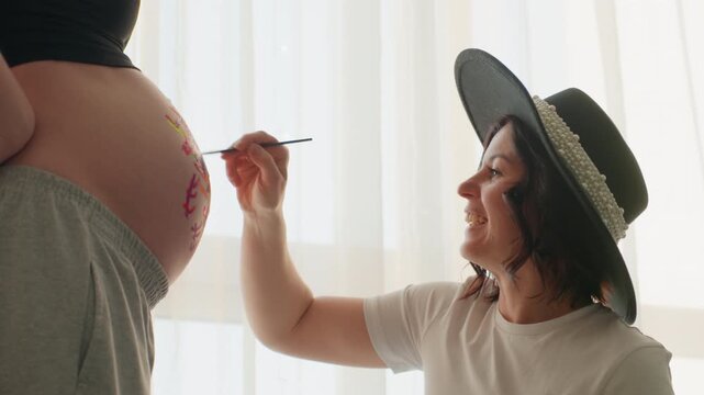 White pregnant woman belly painting in boho home, friend artist in straw hat adds floral brushstrokes, sunlight through curtains, candid joyful smile, intimate maternity ritual, casual wardrobe, warm
