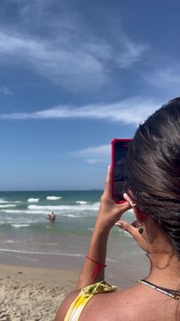 Young woman recording beach vacation on smartphone at sunny ocean shore