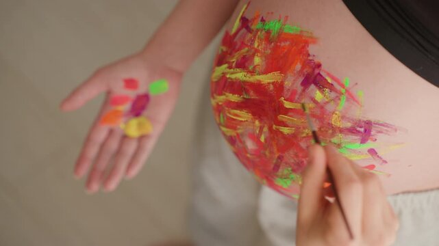 Pregnant belly painted with vibrant abstract strokes, expectant mother holding colorful paint dabs on palm while artist brushes warm acrylic across bump, playful studio light, closeup skin texture