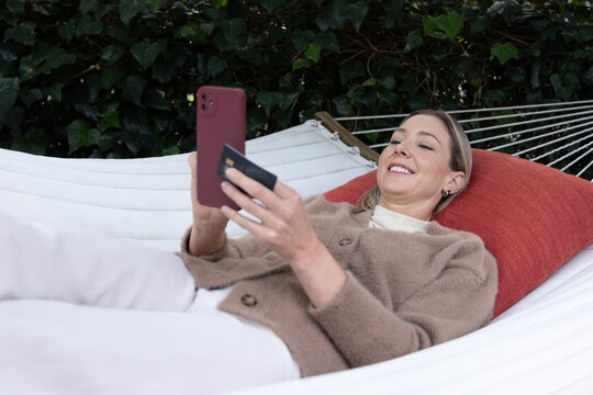 Woman reclining on white hammock holding red phone and card, wearing beige cardigan garden hedge