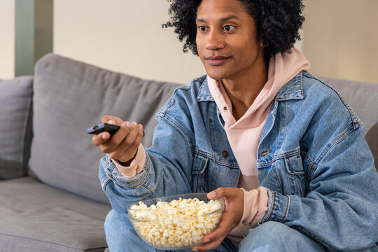 African American man in denim jacket sitting on couch at home holding popcorn bowl pointing remote