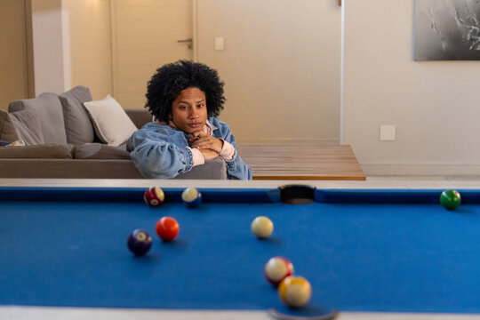 African American man 20s leaning on living room sofa wearing denim jacket watching blue pool table