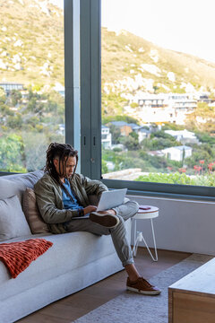 African American man in jacket sitting on gray sofa in living room using laptop, copy space